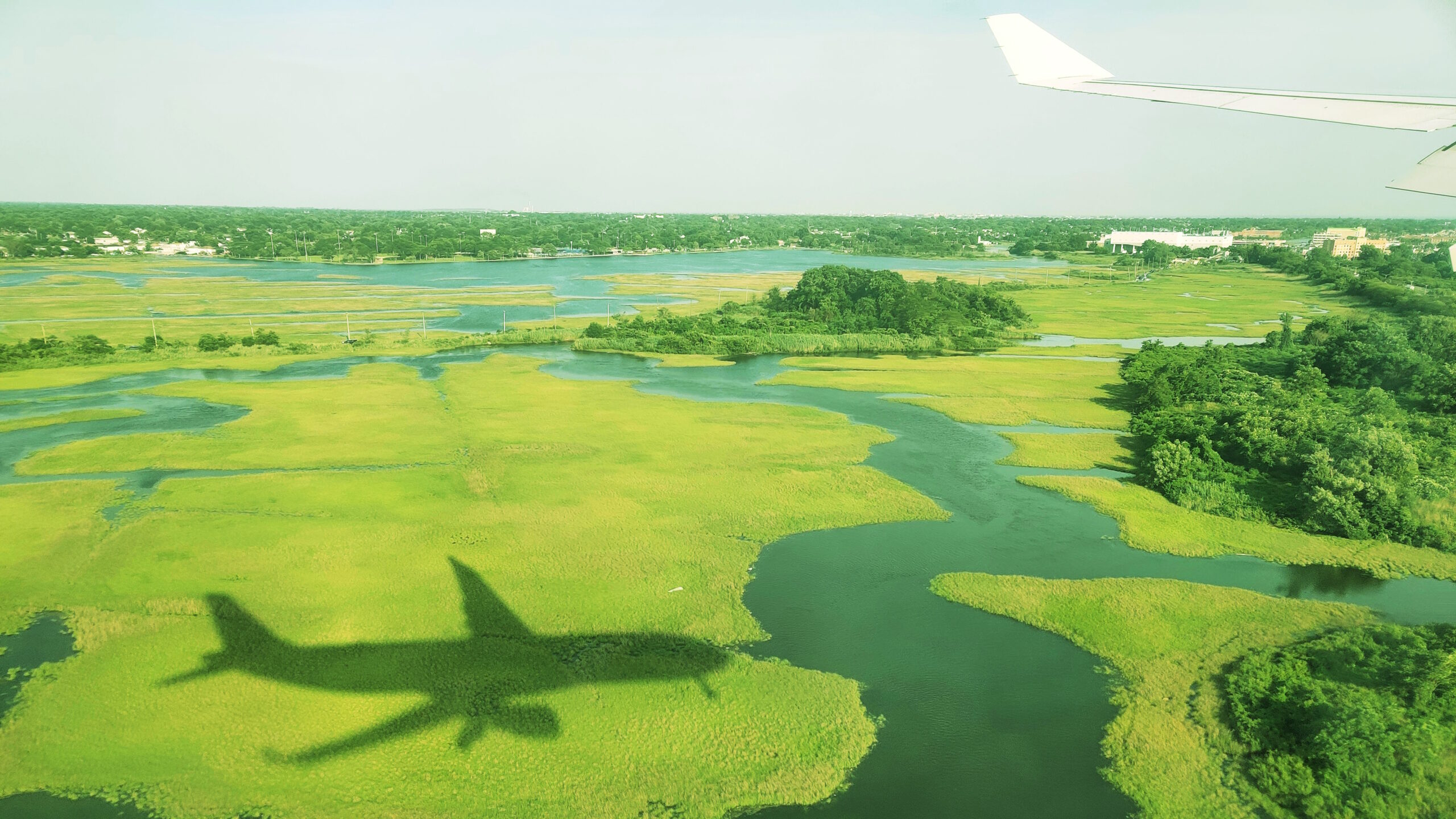[ID: A lush green landscape, with a plane in flight casting a shadow over it. End ID]