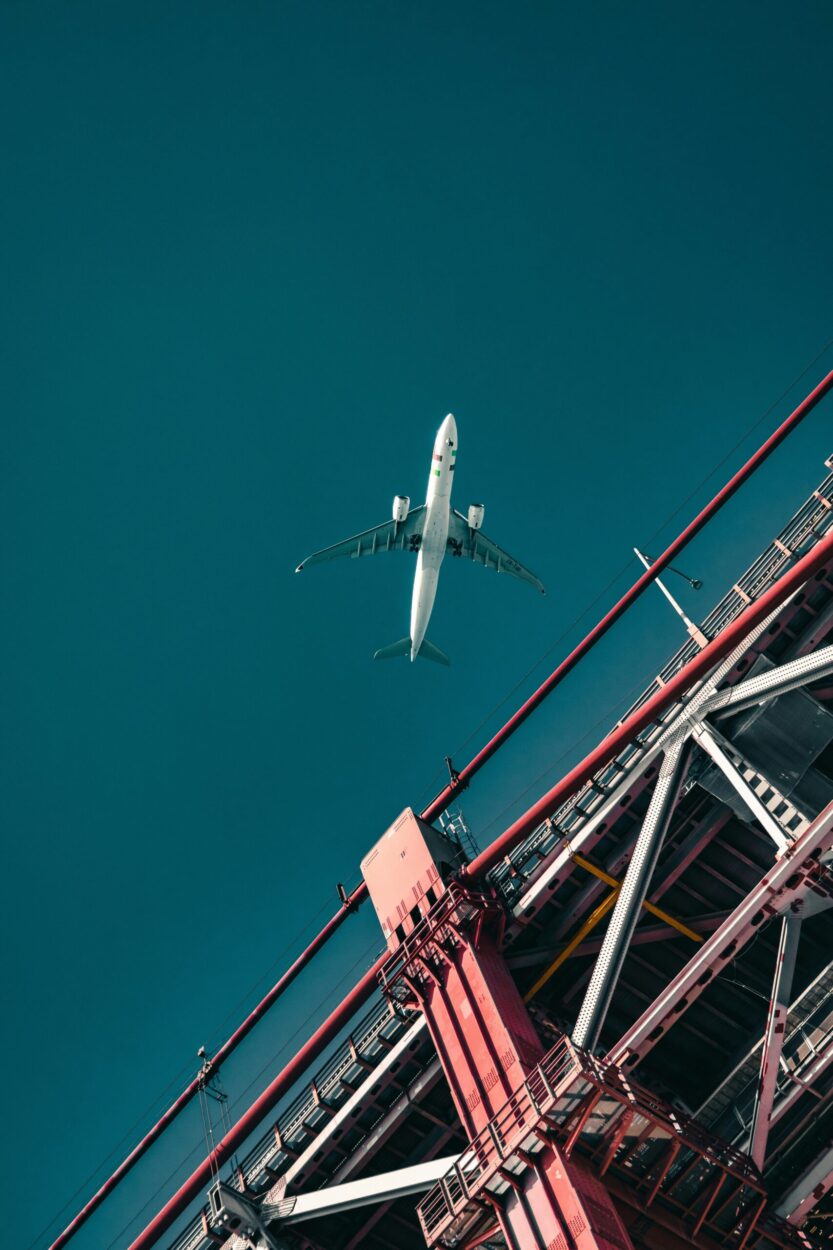 [ID: A plane in flight seen from below against a blue sky. A metal bridge takes up the lower right corner of the image. End ID]